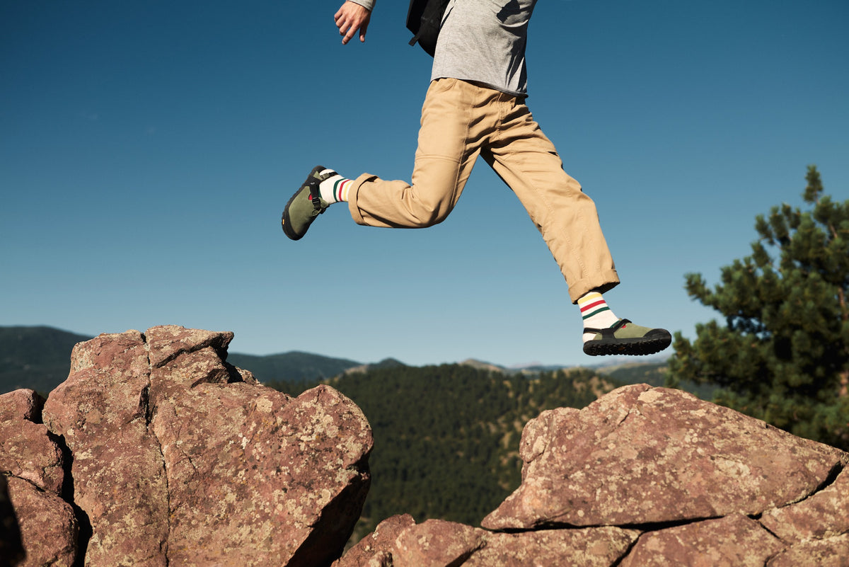 A person jumping between rocks wearing a pair of sagebrush green leather suede mountain clogs