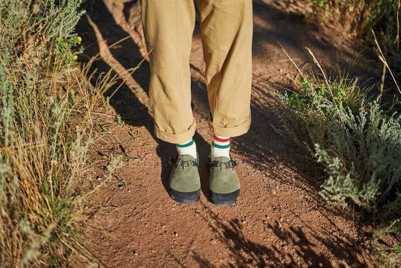 person wearing sagebrush leather suede mountain clogs with red and green striped socks on a trail