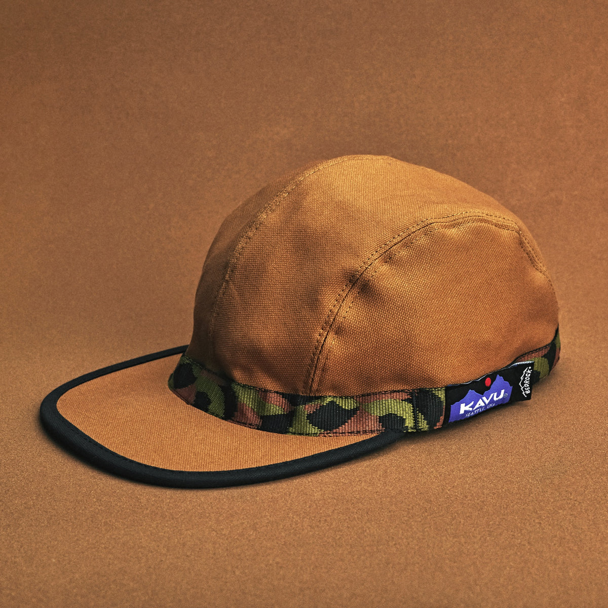 A studio photo with tonal brown backdrop of a brown organic cotton strapback hat with 'KAVU' text on the adjustable strap and a 'Bedrock' patterned webbing visor.