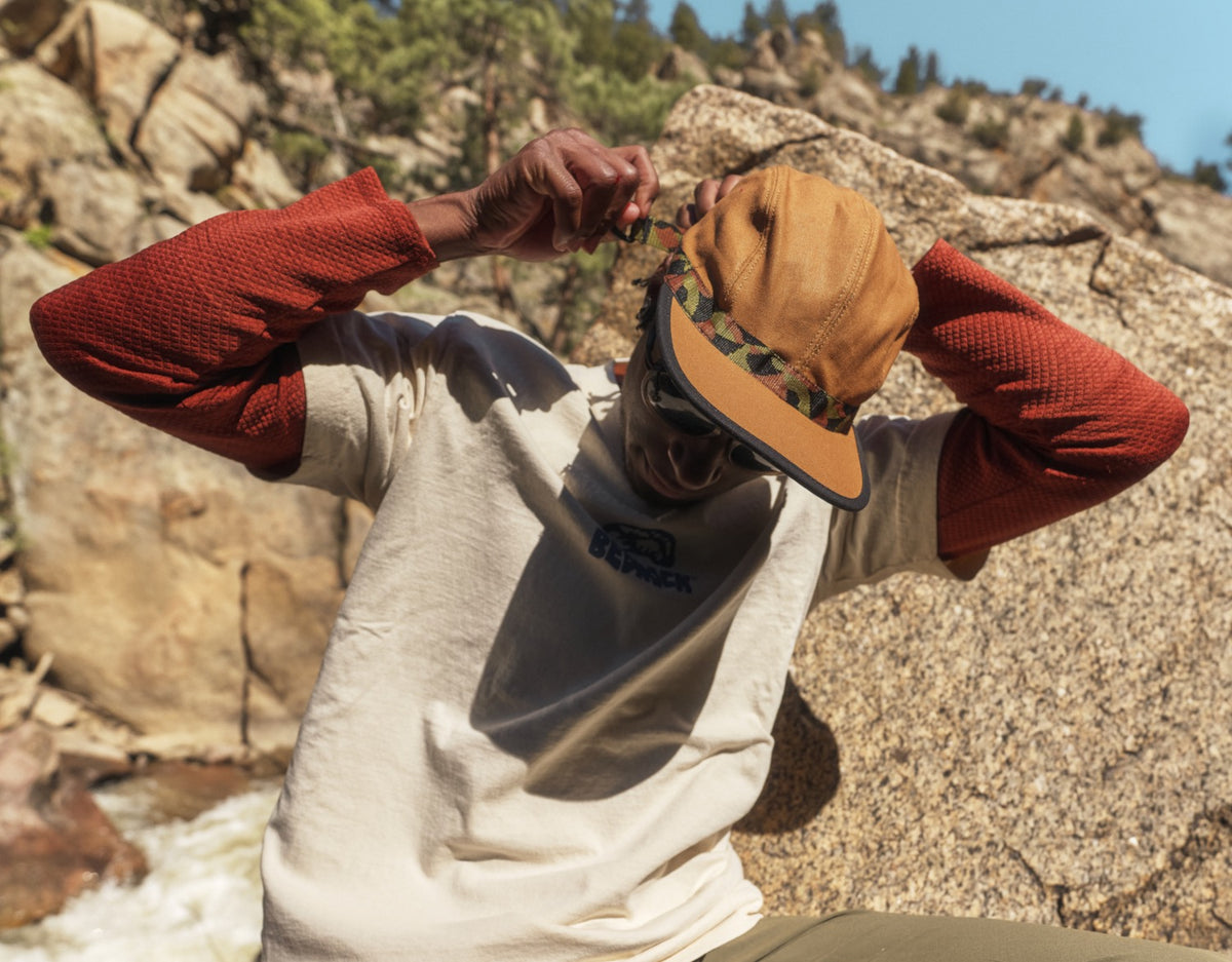 A person looking at the ground wearing a brown organic cotton strapback hat with 'KAVU' text on the adjustable strap and a 'Bedrock' patterned webbing visor.