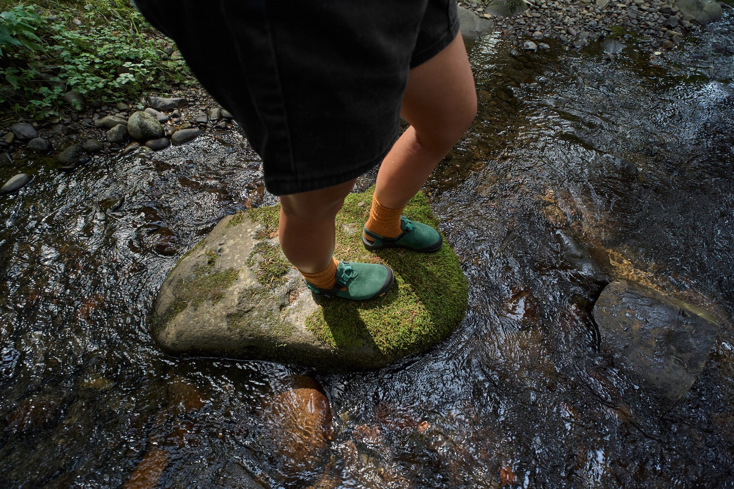 Person stepping onto a moss-covered rock in a natural setting wearing Mountain Clogs in Cascade Green Leather Suede