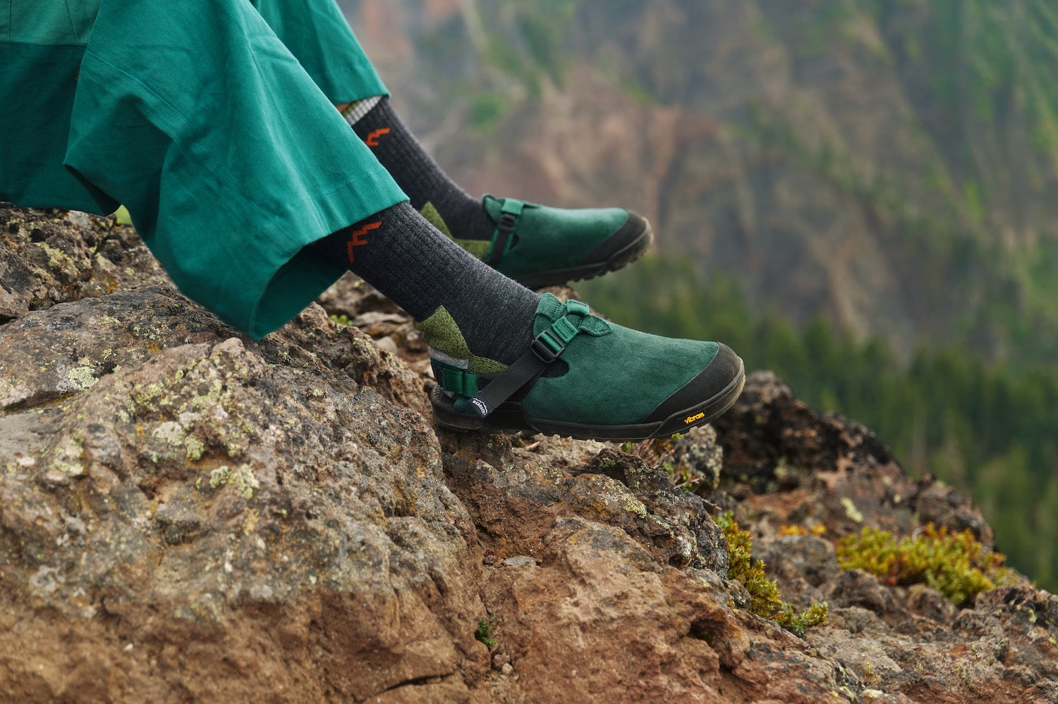 Person wearing Mountain Clogs in Cascade Green Leather Suede and pants sitting on a rocky mountain ledge with a blurred natural background.
