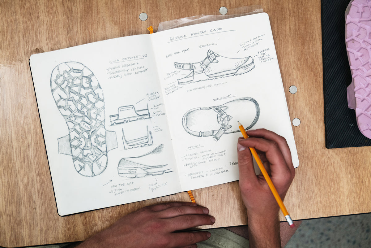 A top-down view of a person's hands sketching shoe designs in a notebook on a wooden desk, with a pink textured shoe sole visible on the right.