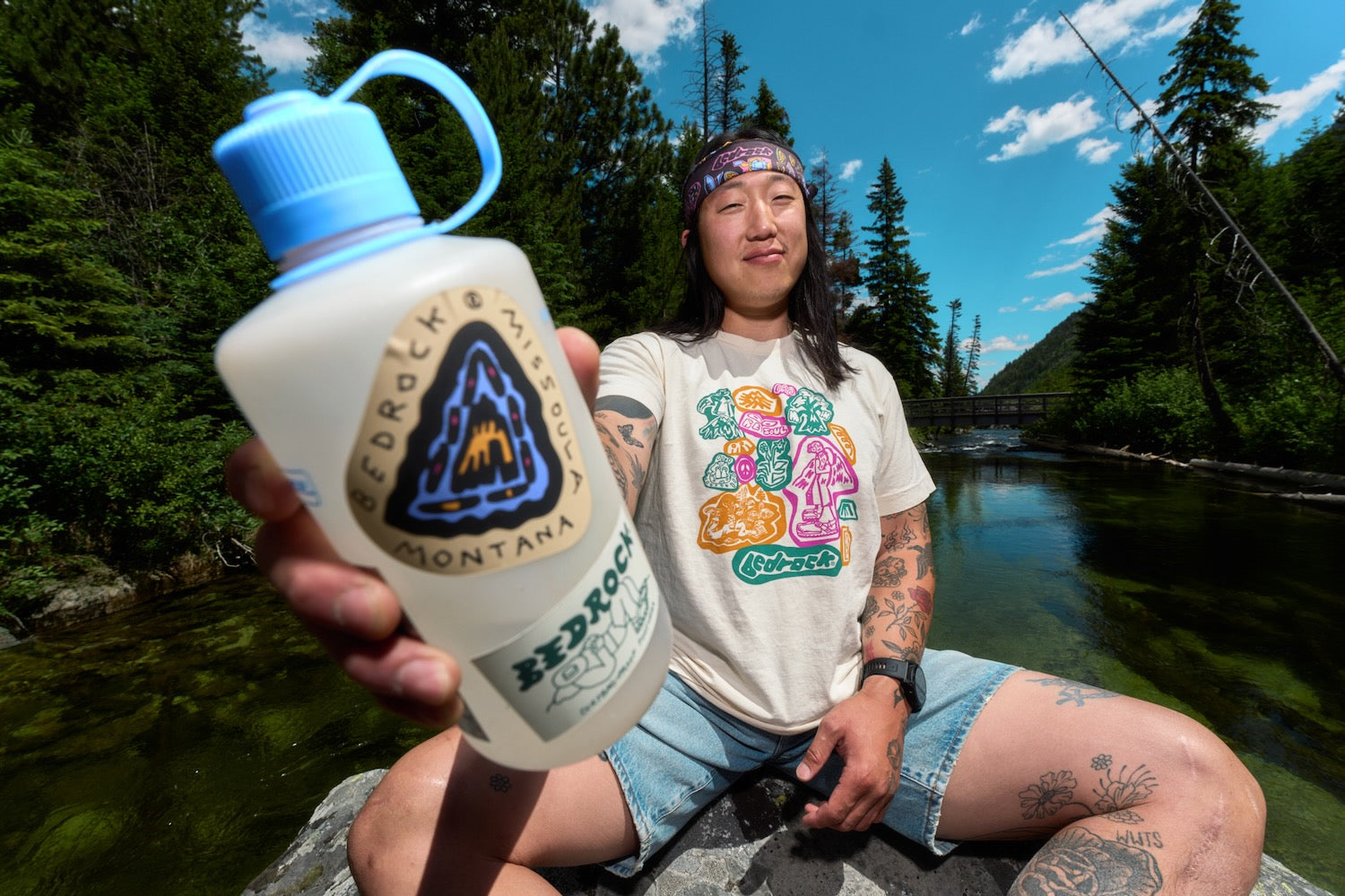 A person sitting outdoors by a river holds up a large white Nalgene water bottle with a blue cap and handle. The bottle has a prominent blue and black mountain logo with a sticker with words 'Bedrock MONTANA' and 'NALGENE' visible.