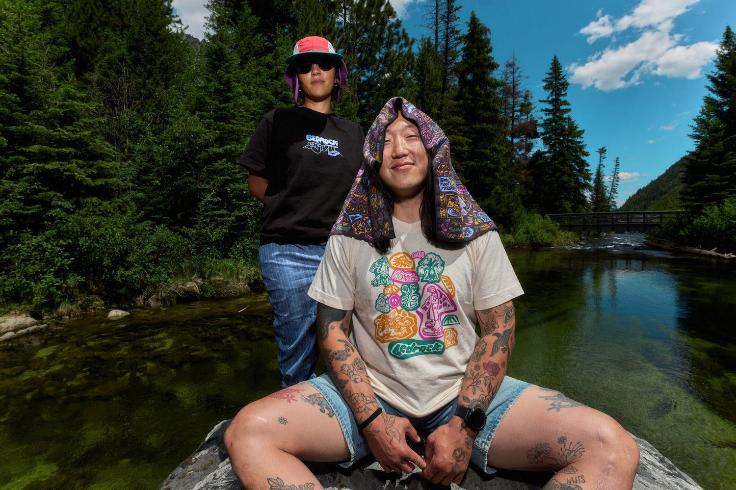 Two people outdoors by a river with a forest and mountains in the background. The seated person in the foreground wears a beige graphic T-shirt, denim shorts, and a colorful Bedrock PackTowl draped over their head.