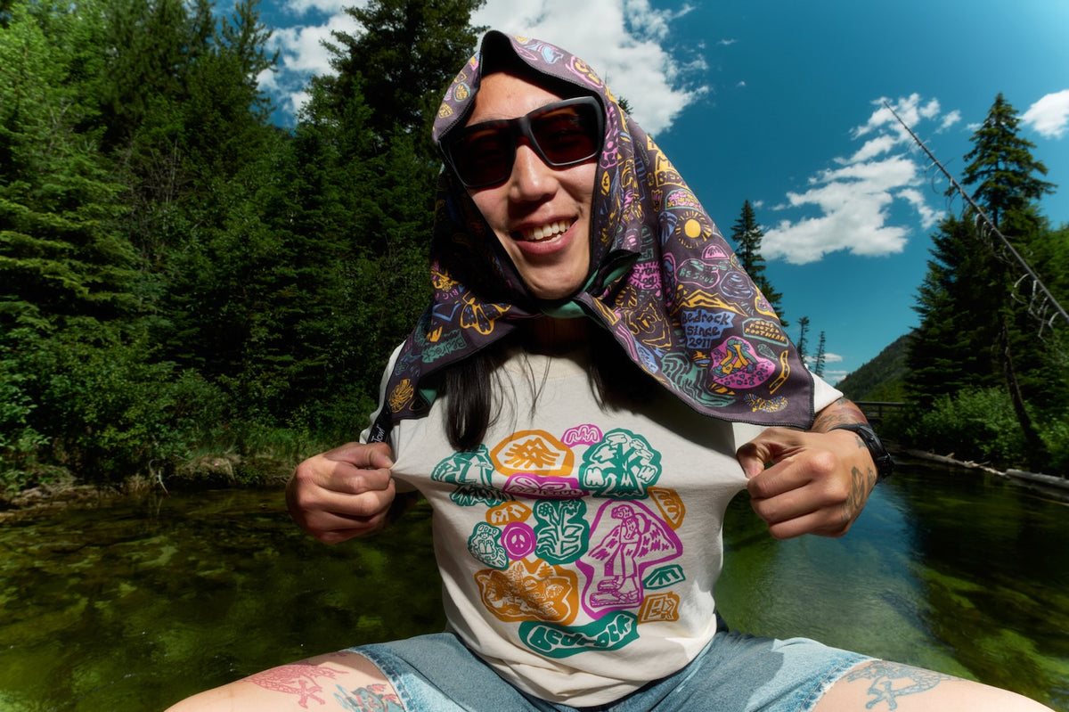 A person outdoors by a river wearing a white graphic T-shirt with a colorful psychedelic mushroom print, a patterned headscarf, and sunglasses, holding the shirt open to show the graphic.