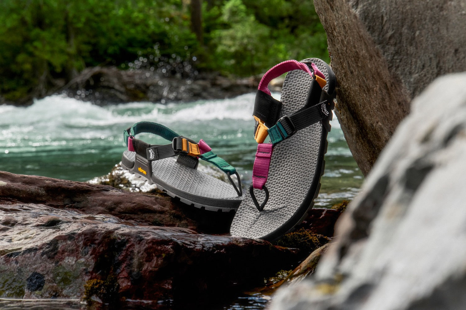 A pair of Bedrock metamorphic hiking sandals resting on wet rocks beside a rushing river, with a forest in the background. The sandals feature textured black and colorful straps in black, magenta, teal, and orange.