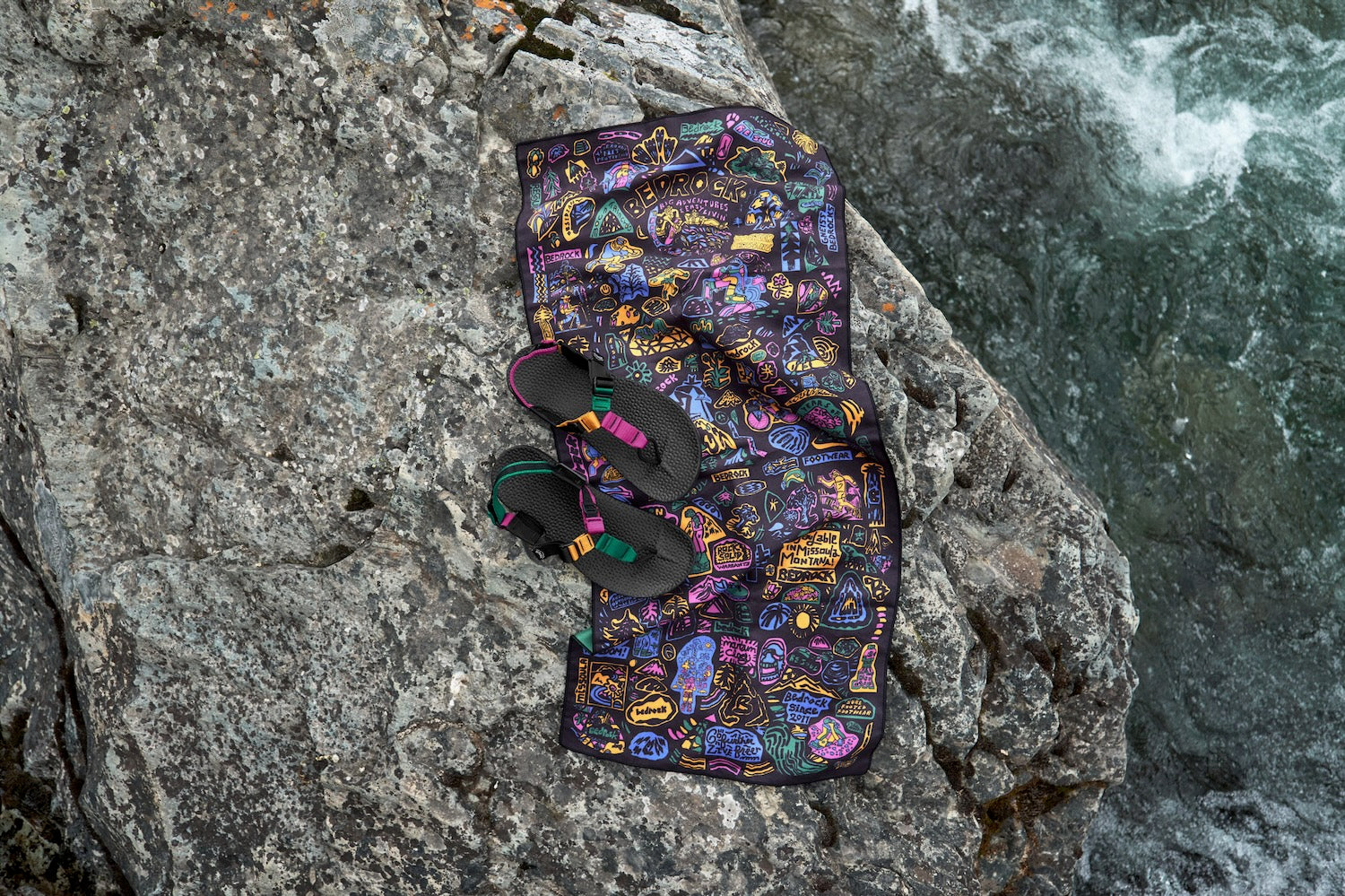A colorful Bedrock Archive PackTowl and a pair of Bedrock Sandals with bright straps are arranged on a large gray rock beside rushing water.
