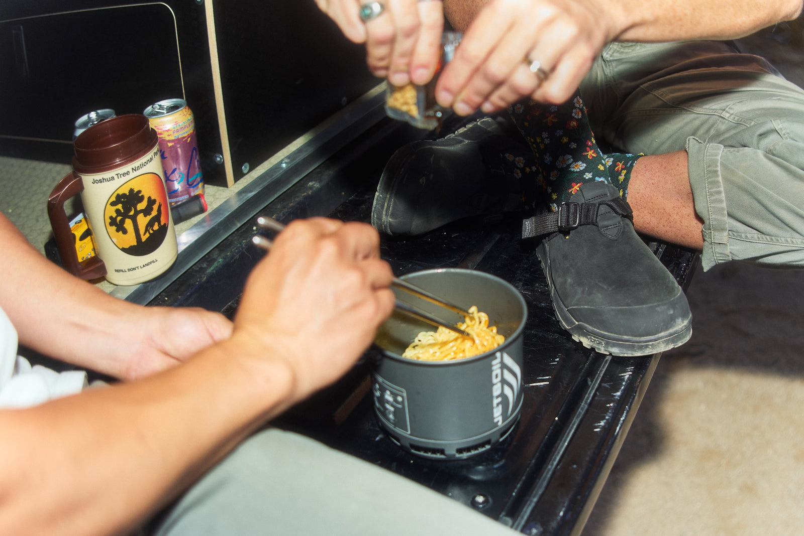 Two people preparing food on a camping stove with various items around wearing obsidian dark gray nubuck leather mountain clogs
