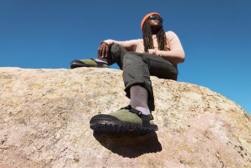 person sitting on a rock wearing sagebrush green leather suede mountain clogs with blue sky background