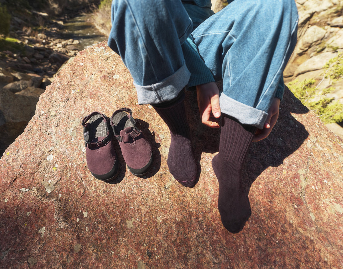 Person posing on a rock wearing purple socks about to put on Deep Plum Mountain Clogs