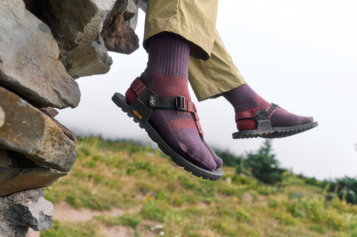 Person wearing purple sandals and Crew length plum split-toe socks standing on rocks with a natural landscape in the background