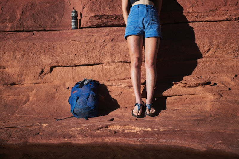 Person wearing a pair of cairn evo 3d sandals with slate blue straps standing in front of red desert rock wall