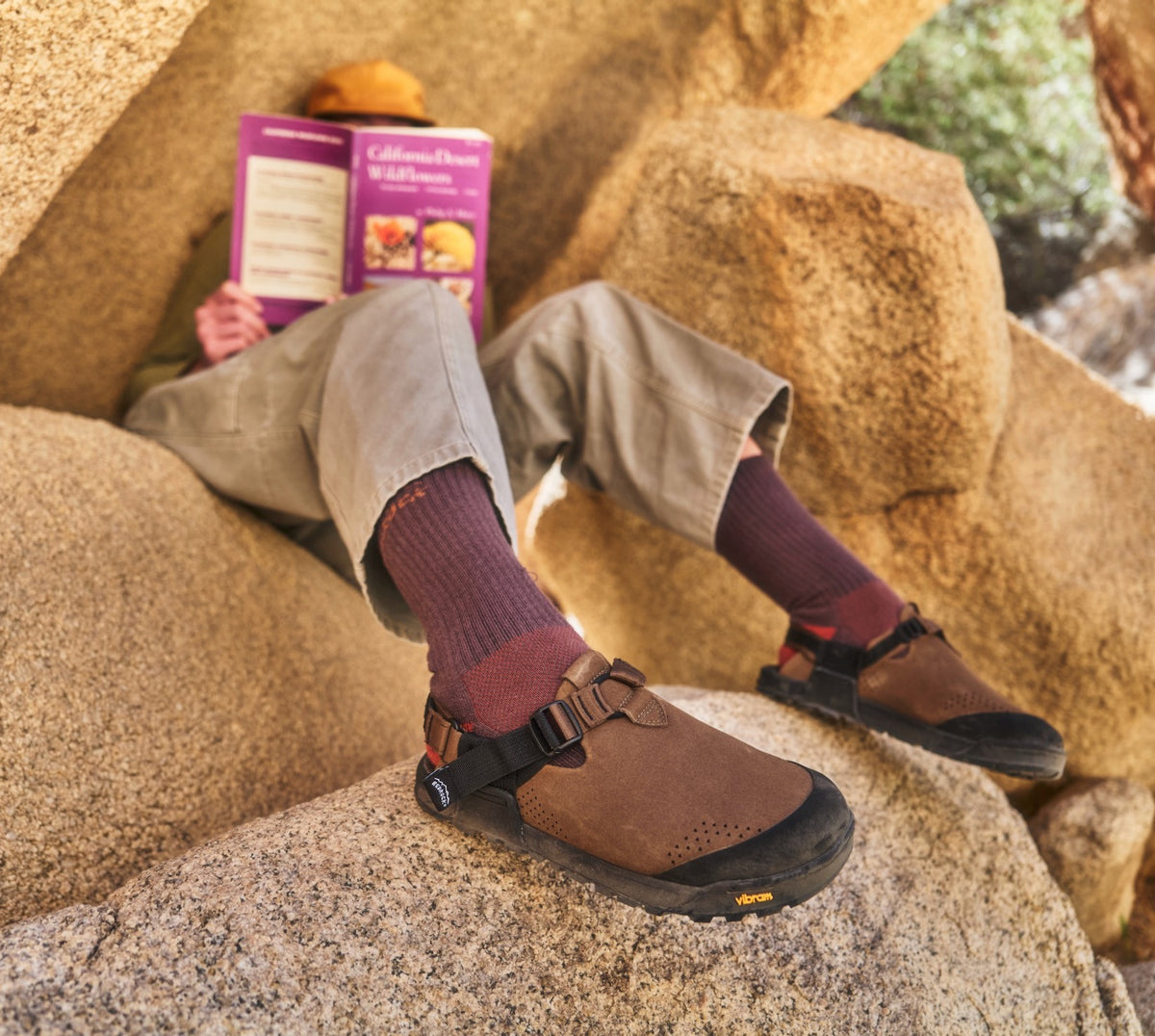 Person wearing Pinecone Synthetic Suede Mountain Clogs  and purple socks sitting on rocks with a book.