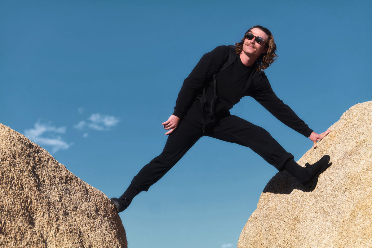 Person in black outfit standing on a rock with a clear blue sky, wearing Obsidian Black Synthetic Mountain Clogs 