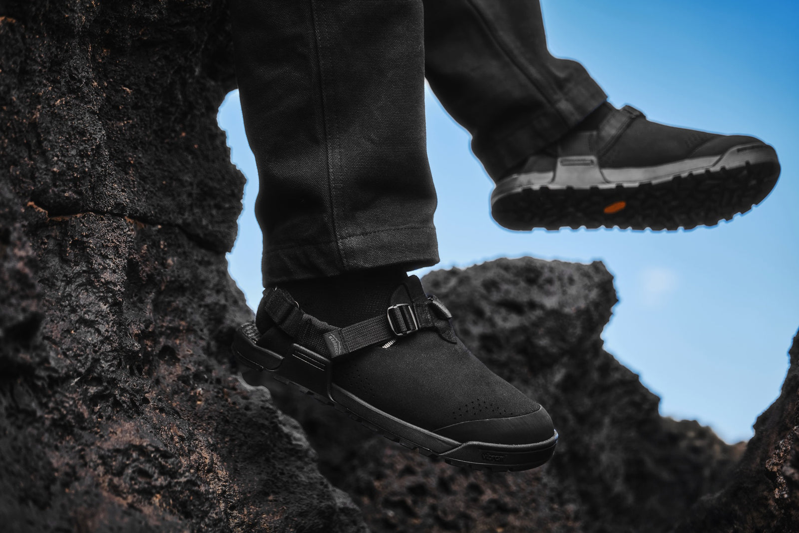Person wearing obsidian black synthetic suede clogs while sitting on a volcanic rock
