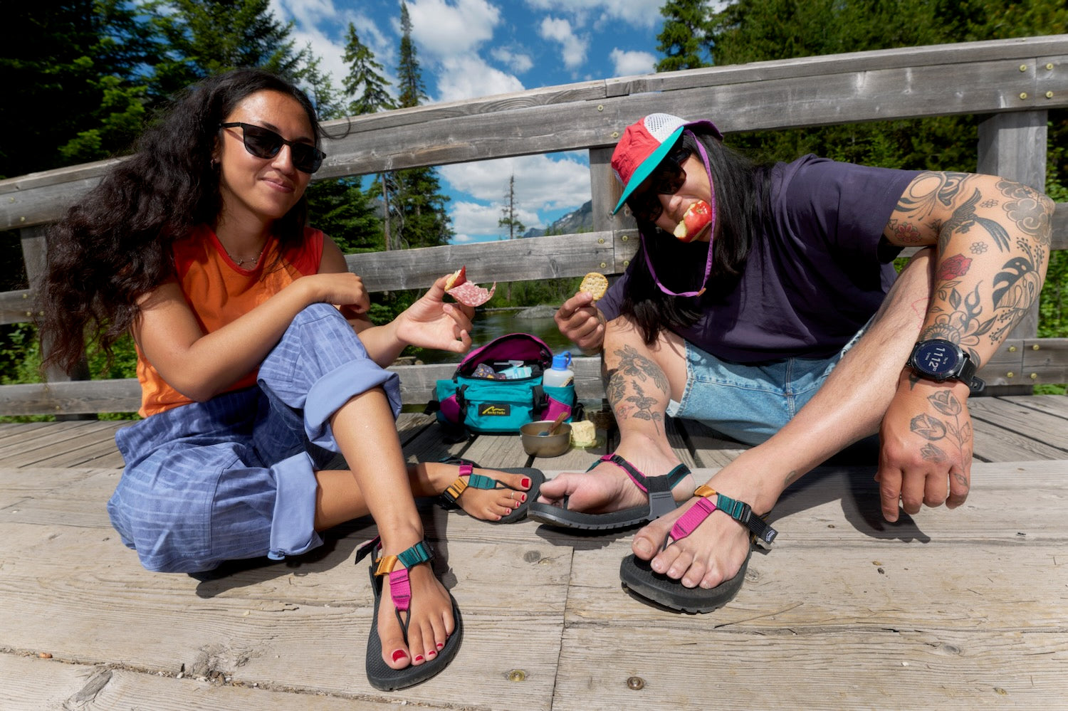 Two people sitting on a wooden deck outdoors, sharing a snack. One person wears an orange shirt and blue jeans, and the other wears a dark purple shirt, denim shorts, and a red cap.