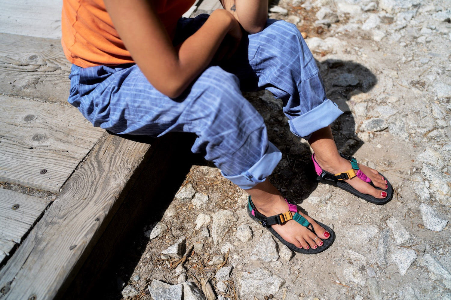 A person sitting outdoors on a wooden bench wearing blue plaid pants, an orange shirt, and black sandals with A pair of Bedrock Cairn Metamorphic sandals.