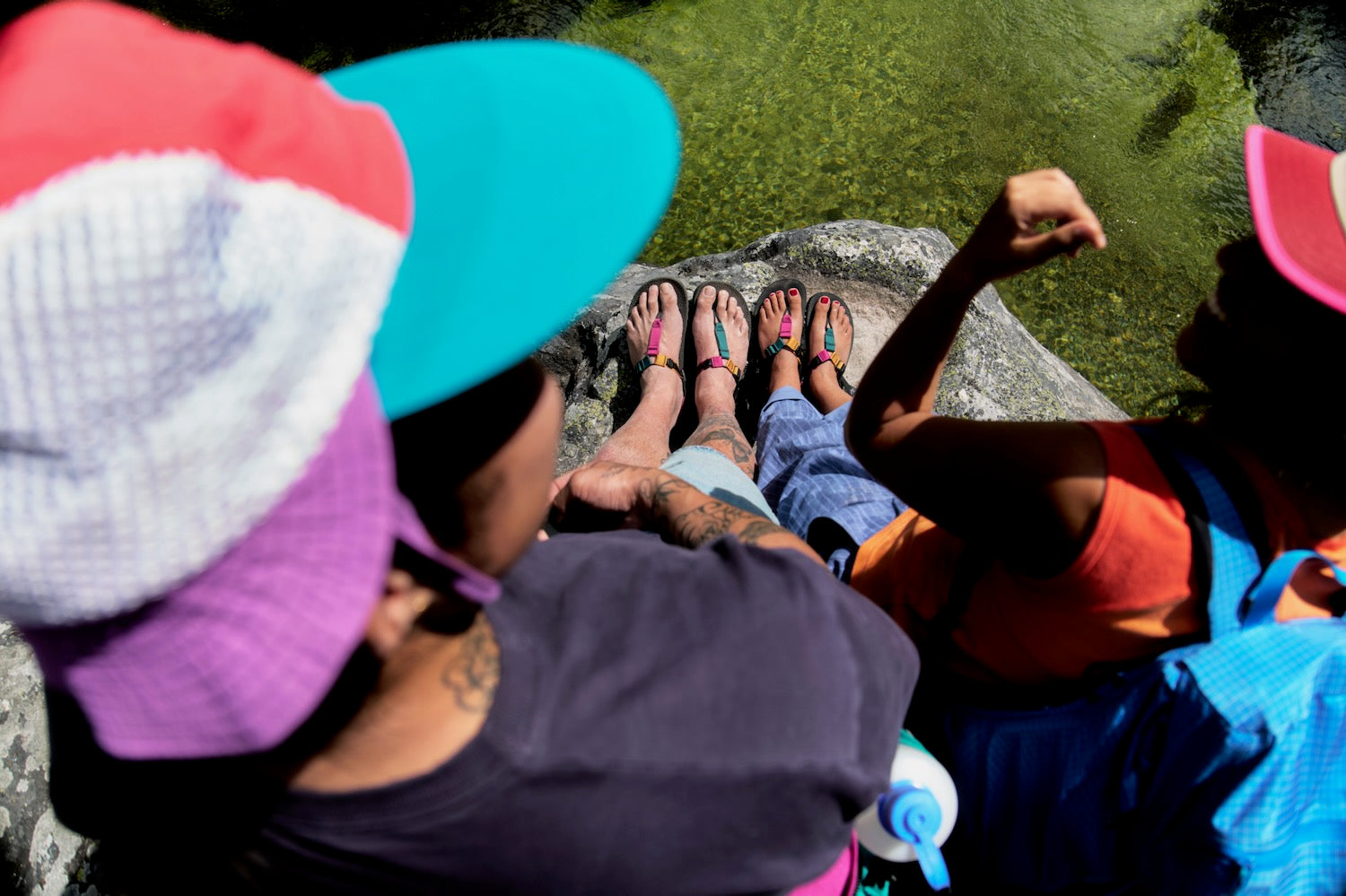 Several people sitting outdoors on a rock by the water, wearing colorful hats and sandals. The scene is viewed from above, with the heads and legs of the group visible against green water and a gray rock.
