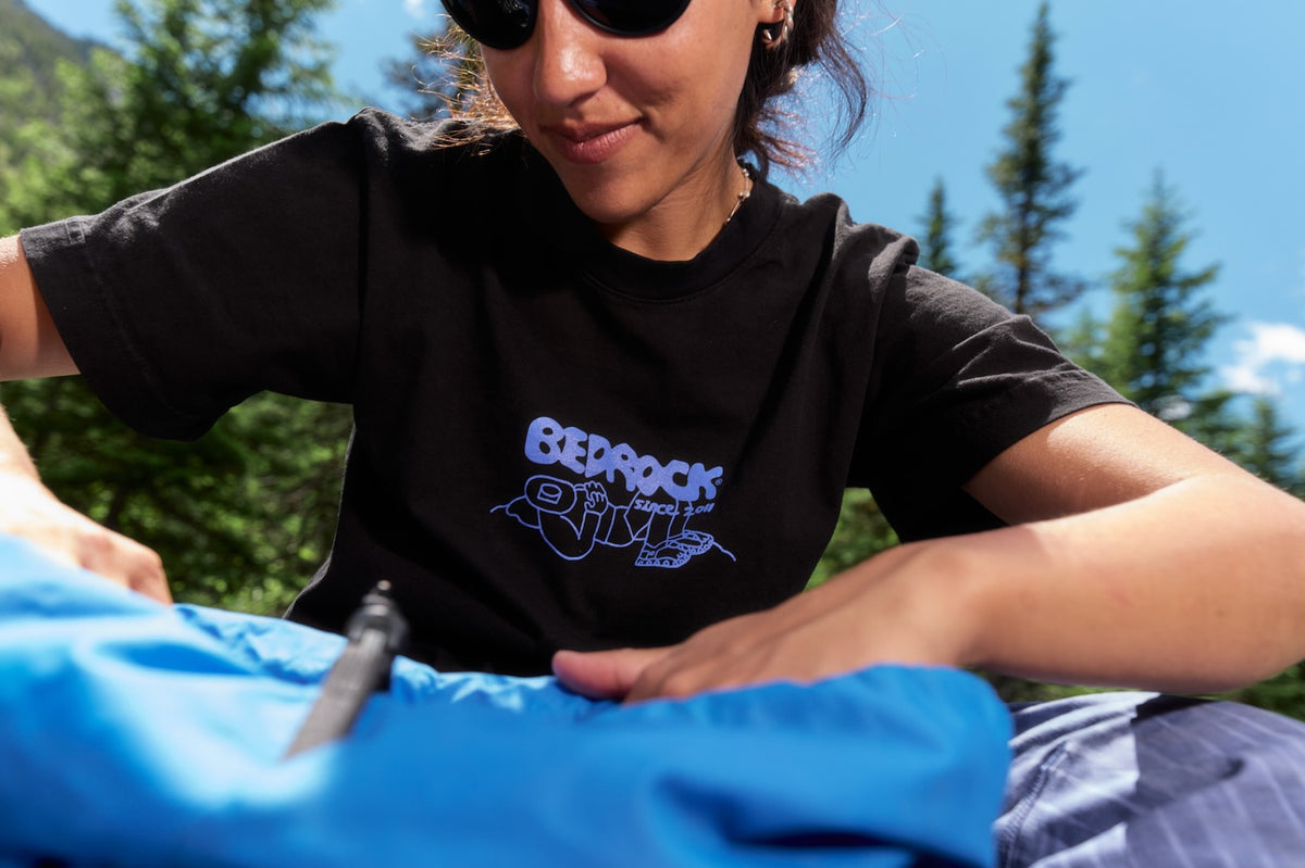 A woman wearing a black t-shirt with a blue 'Bedrock logo, sitting outdoors in a sunny forest setting and handling a bright blue diving mask.