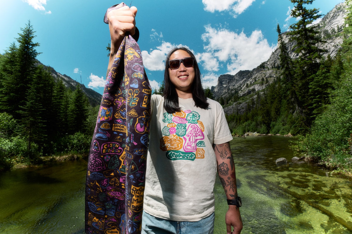 A man wearing sunglasses and a graphic t-shirt stands outdoors in front of a mountain river landscape, holding up a long patterned scarf or shawl with colorful abstract designs.