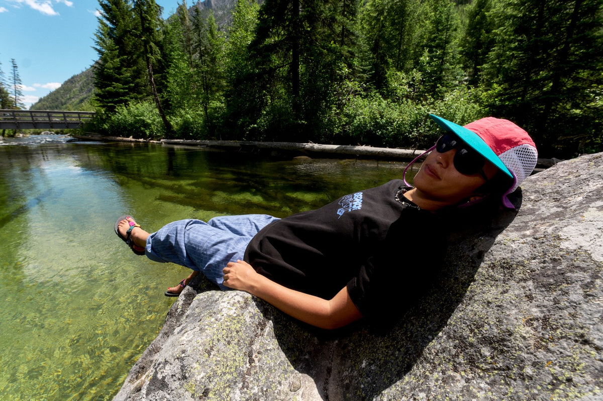 A person wearing a black Bedrock t-shirt, blue jeans, sunglasses, and a pink and teal cap relaxes on a large rock beside a clear mountain stream with a forest in the background.