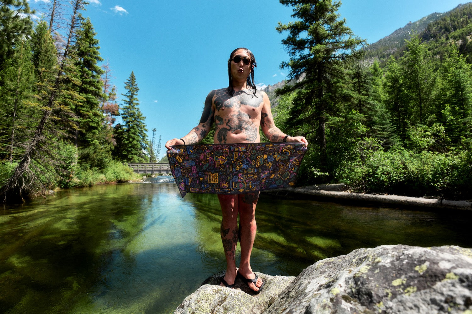 A tattooed man wearing sunglasses stands on a rock by a river, holding up a colorful, patterned rectangular Bedrock PackTowl. The background features a lush green forest, mountains, and a clear blue sky.