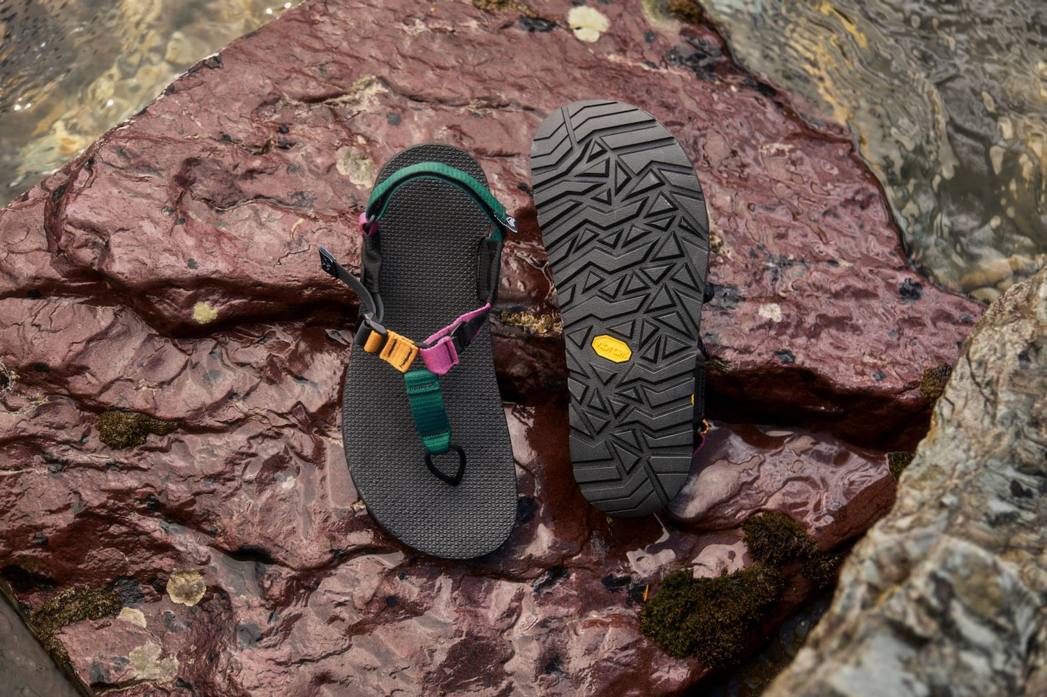A pair of Bedrock Cairn Metamorphic sandals shown on wet reddish-brown rocks outdoors. One sandal is placed upright to show the black footbed and colorful straps, and the other is turned to show the treaded outsole with a small yellow Vibram logo.