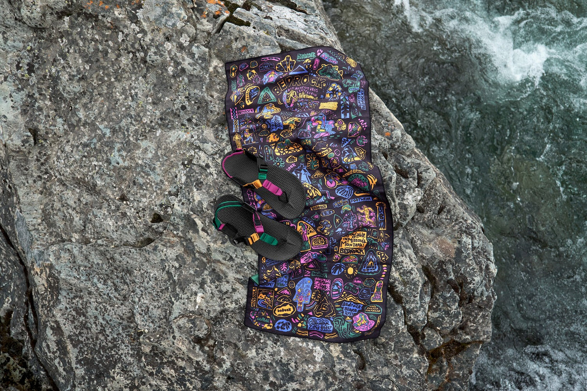 A colorful Bedrock Archive PackTowl and a pair of Bedrock Sandals with bright straps are arranged on a large gray rock beside rushing water.