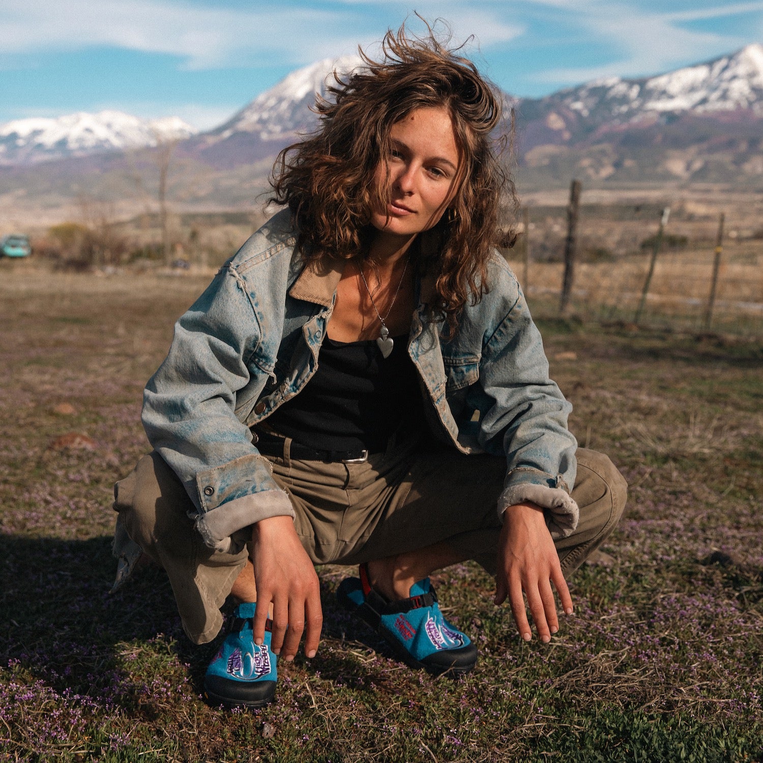 Person squatting in a field with mountains in the background wearing Stone Wizard Cyber Blue Clogs 